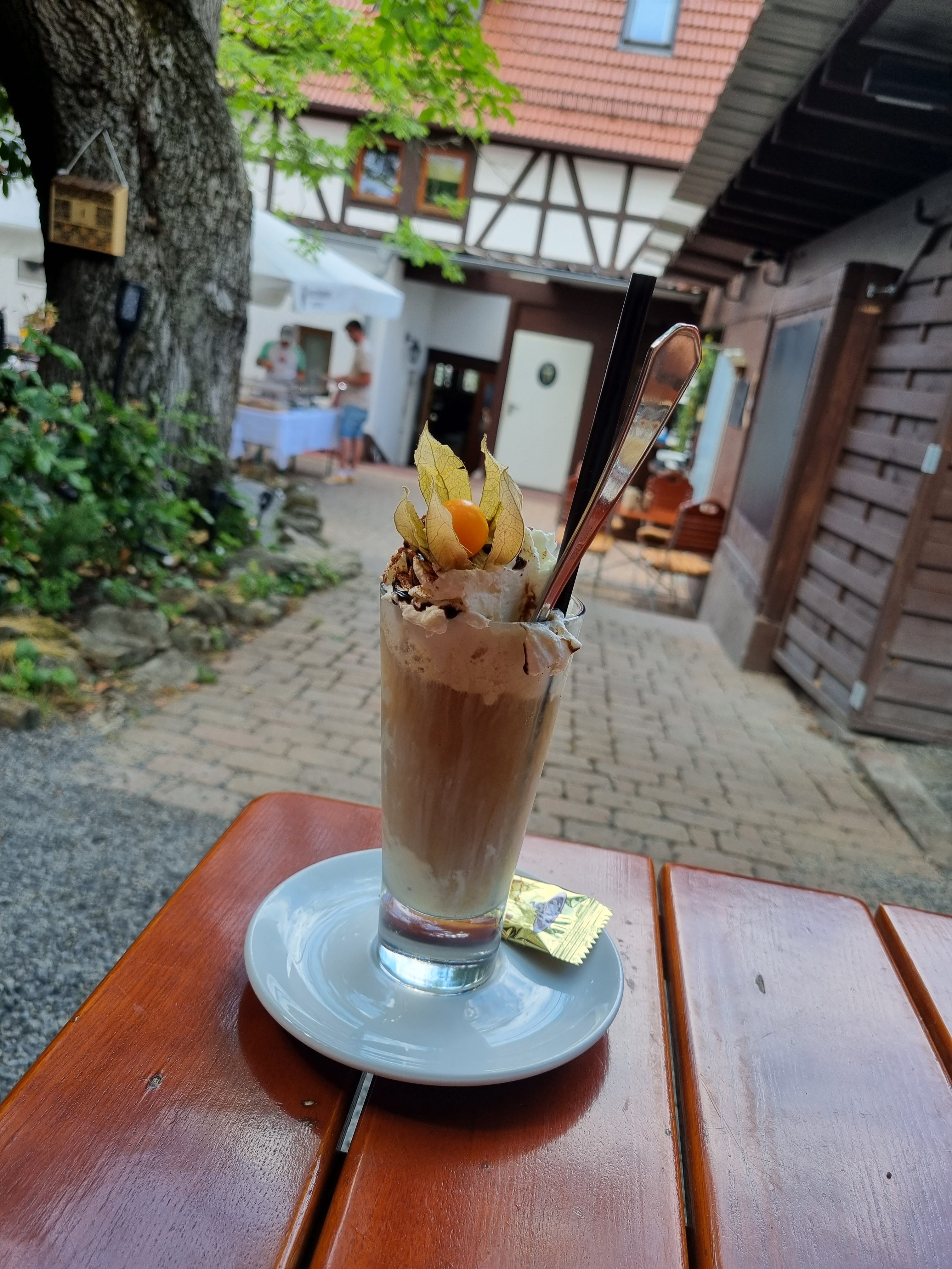 Ein Eiscafé-Getränk in einem hohen Glas steht auf einem Holztisch im Freien. Das Getränk ist mit Sahne, Schokoladenraspeln und einer Physalis-Frucht garniert. Daneben liegt ein kleiner Keks. Im Hintergrund sind ein gepflasterter Weg und Gebäude zu sehen.