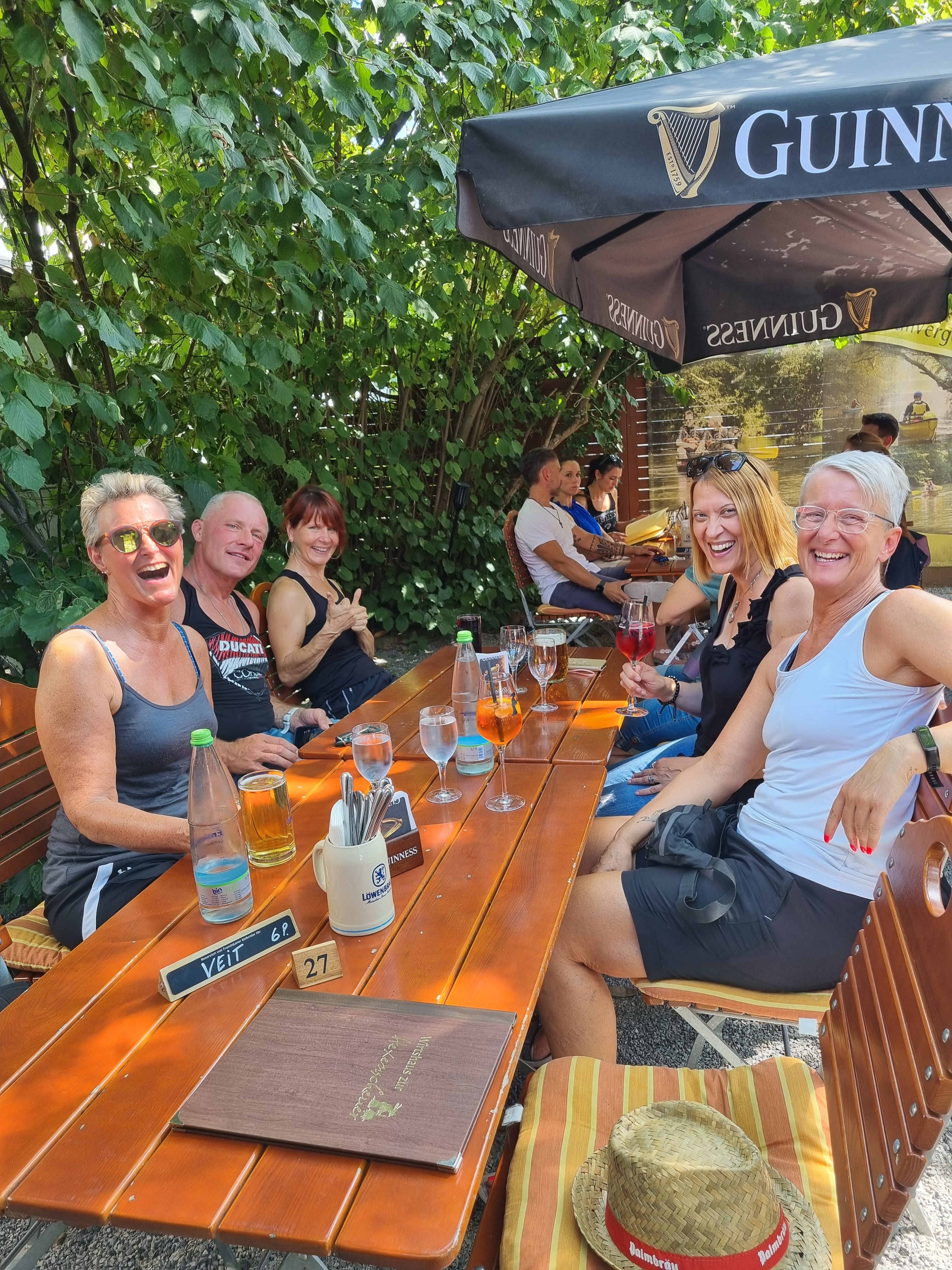 Ein Foto zeigt eine Gruppe von sechs Personen, die an einem langen Holztisch im Freien sitzen. Sie lachen und scheinen sich zu unterhalten. Auf dem Tisch stehen Getränke und Gläser. Im Hintergrund sind grüne Pflanzen und ein Sonnenschirm zu sehen.
