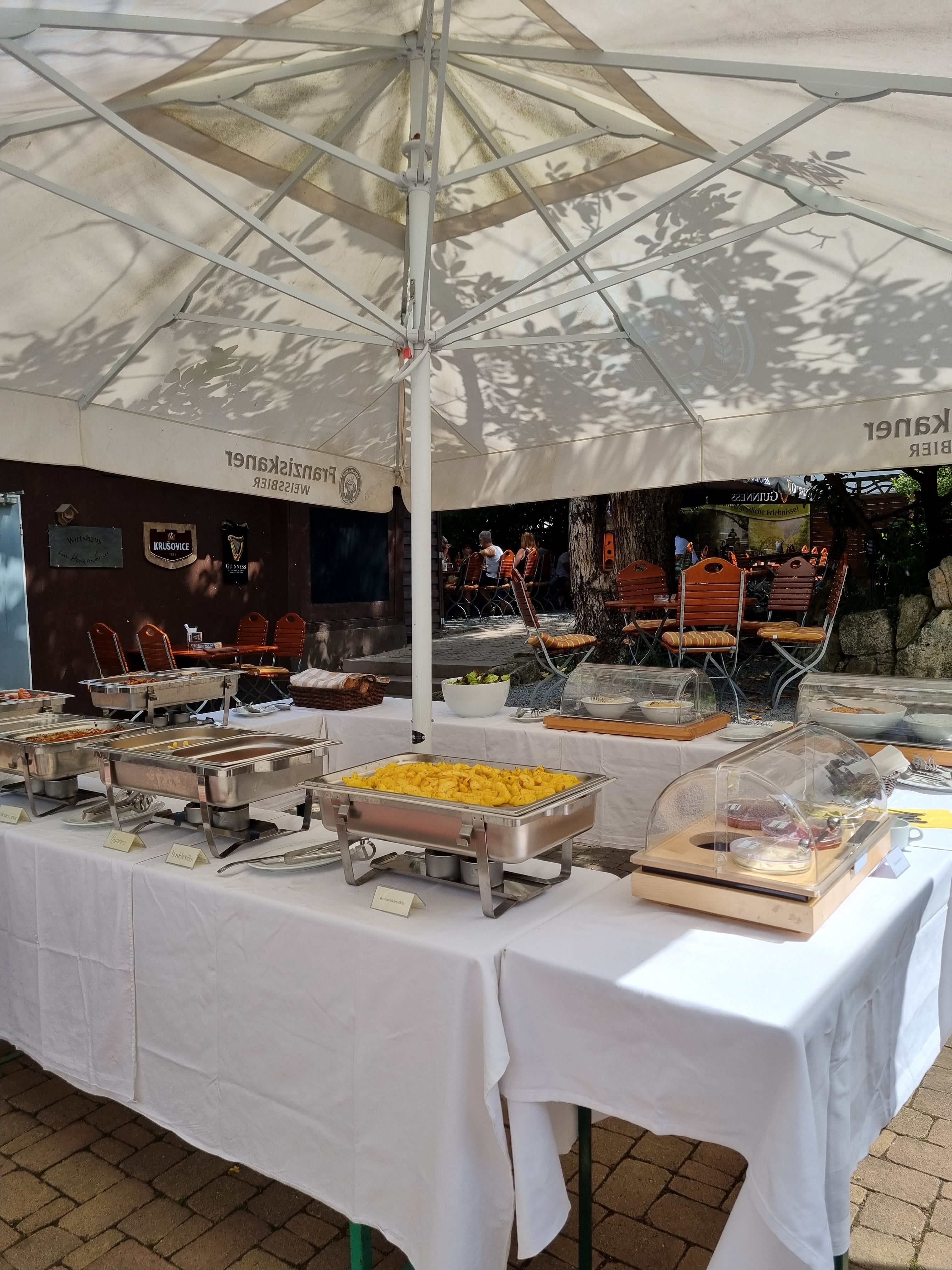 Ein Buffet-Tisch unter einem großen, weißen Sonnenschirm im Freien. Auf dem Tisch stehen mehrere silberne Warmhaltebehälter mit Speisen. Dahinter sind Stühle und einige Personen im Hintergrund zu sehen.