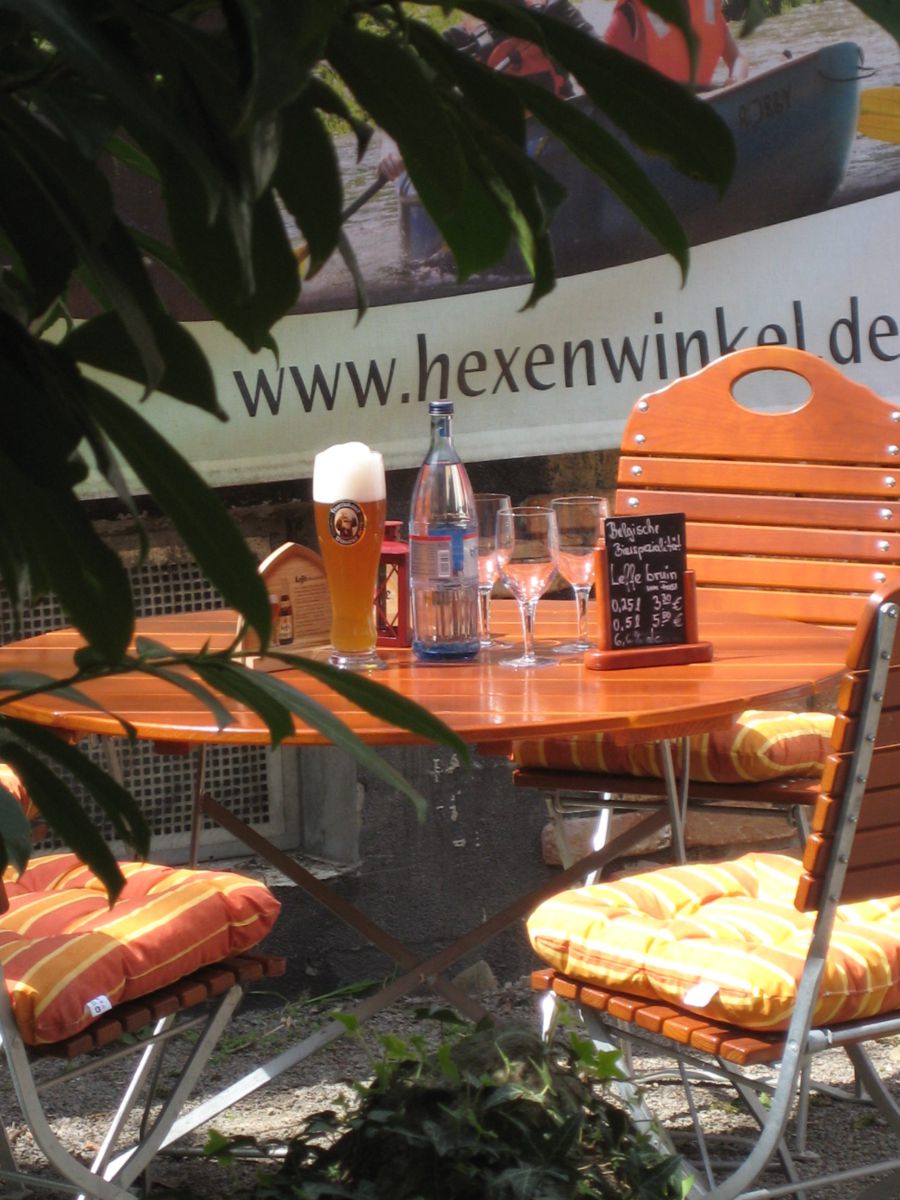 Ein runder Holztisch im Freien ist gedeckt mit einem Bier, einer Wasserflasche, zwei Weingläsern und einer kleinen Tafel mit Speisekarte. Auf dem Tisch liegen orange-gelbe Sitzkissen. Im Hintergrund ist ein Banner mit der Aufschrift „www.hexenwinkel.de“ zu sehen.