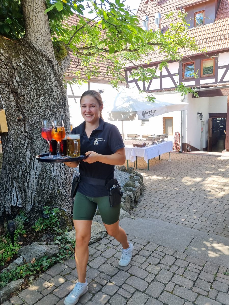 Eine Frau mit dunklem Oberteil und grünen Shorts trägt ein Tablett mit Getränken durch einen Außenbereich. Im Hintergrund sind ein großer Baum, Gebäude mit einem weißen Sonnenschirm und Tische zu sehen. Der Boden ist mit Pflastersteinen bedeckt.