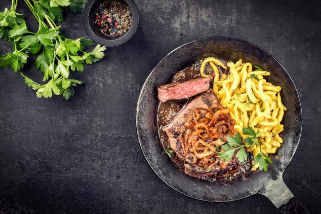 schwarzer hintergrund gusseiserne Pfanne mit rumpfsteak zwiebeln und schwäbische spätzle angerichtet